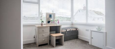 A corner bedroom with a desk, a small stool, a chest of drawers, a radiator, and a window with blinds.