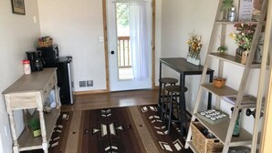 A cozy room with a wooden floor, a small table, a bar stool, a shelf with decorative items, and a door leading outside. - Cabin #1 - Backyard Bunkhouse

 (Irwin)