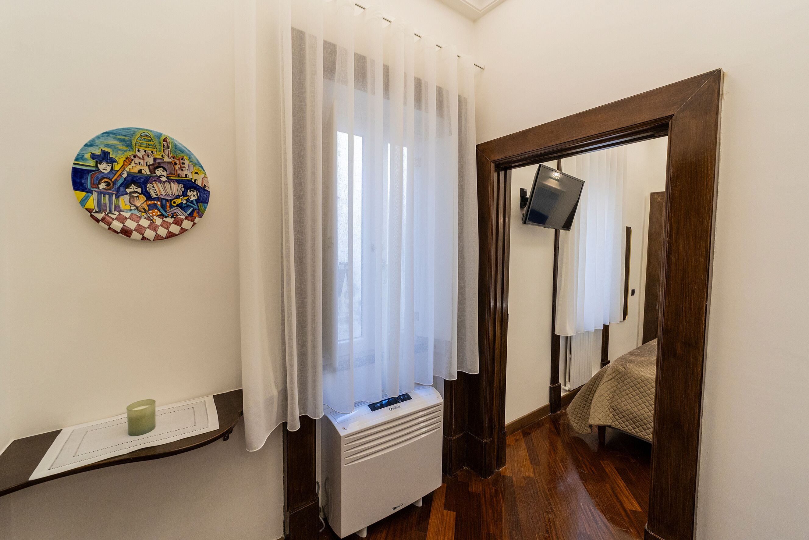 Ein Hotelzimmer mit Bett, Wandfernseher, Heizung, Fenster mit transparenten Vorhängen und einem Wandbild.