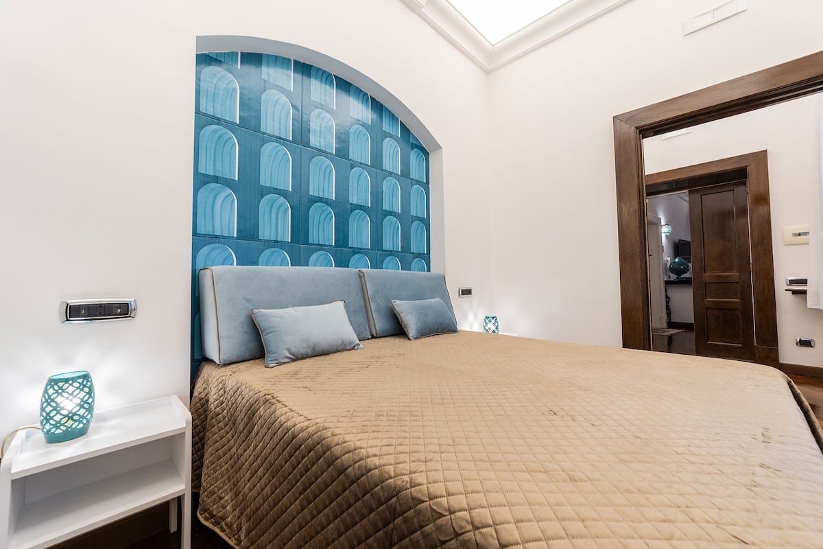 A bedroom with a large bed, a nightstand, a wall-mounted power outlet, and a door to another room.