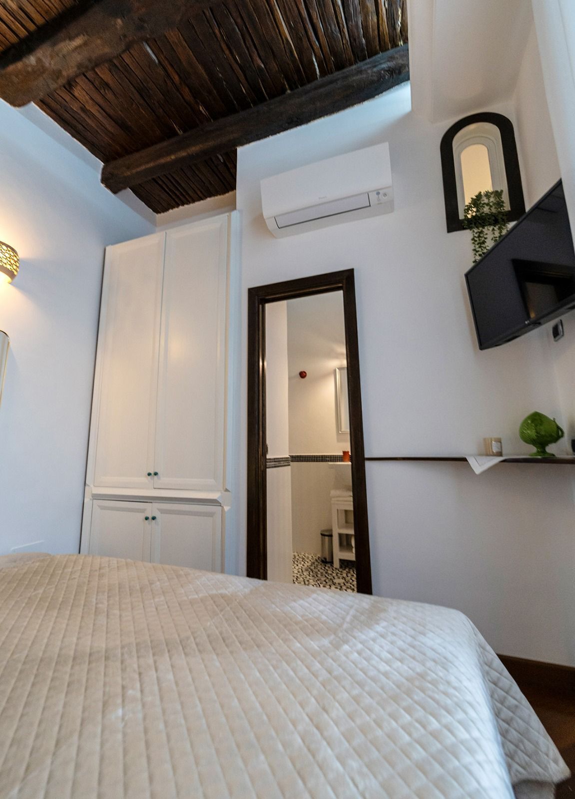 A bedroom with a bed, wardrobe, mirror, TV, and a small plant on a shelf.
