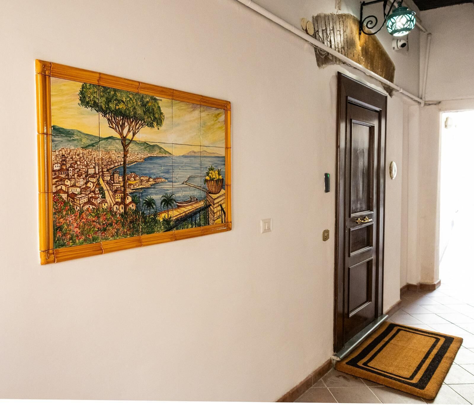 Un couloir doté d’un sol carrelé, une porte avec une poignée en laiton, un luminaire fixé au mur et un grand tableau encadré représentant une scène d’une ville côtière.
