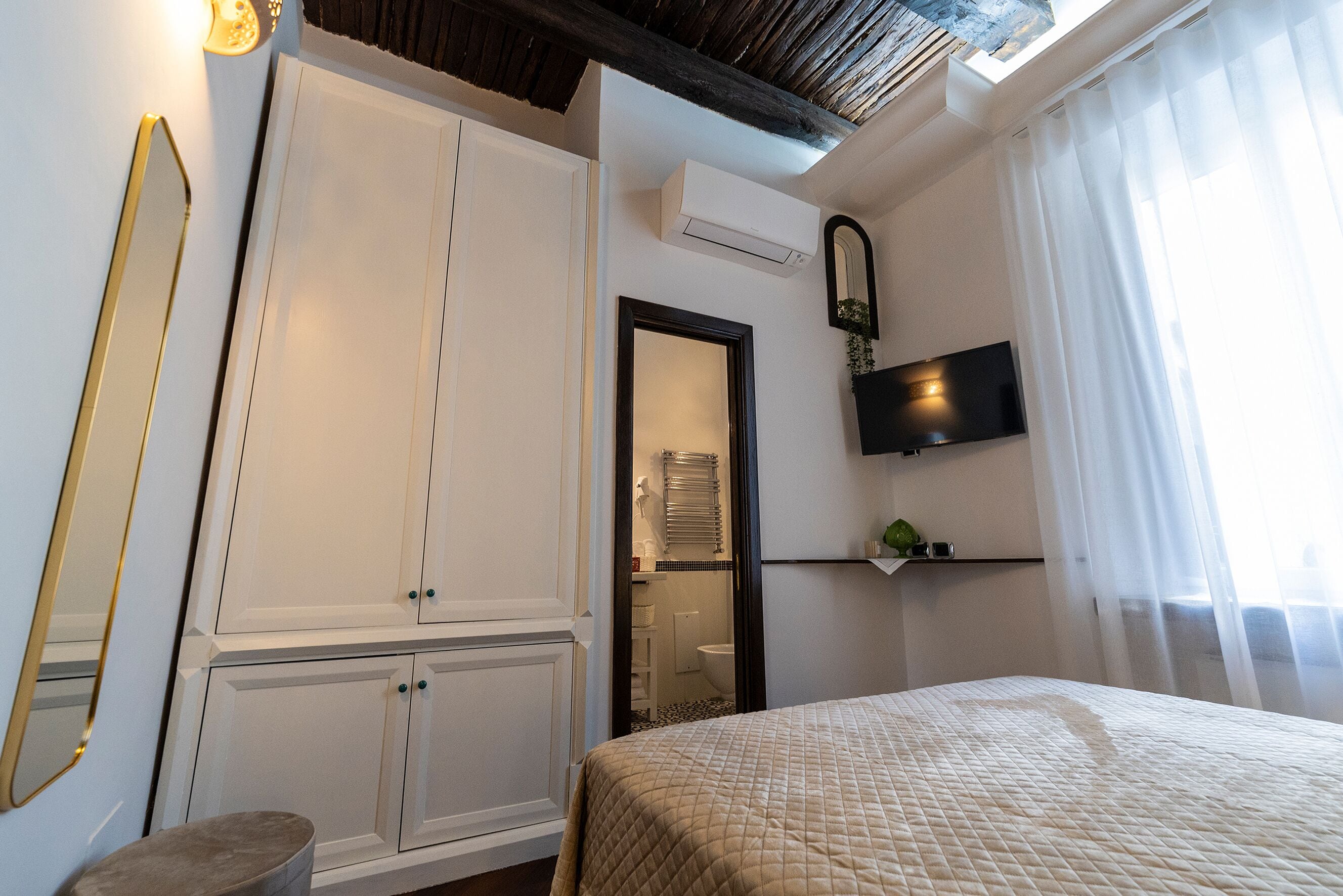 A bedroom with a bed, a wardrobe, a mirror, a television, and a window with curtains.
