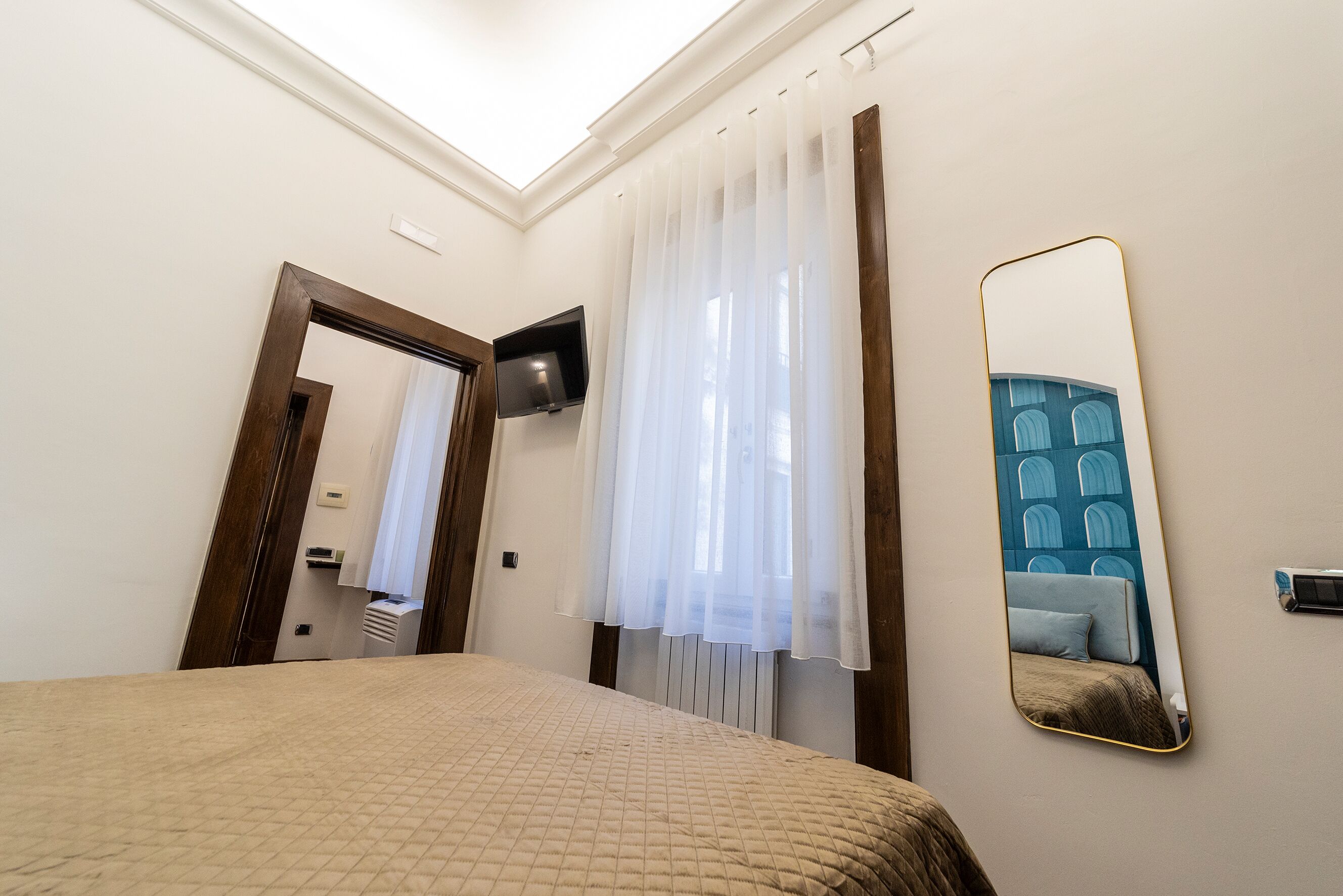Habitación de hotel con cama, televisión montada en la pared, espejo y ventana con cortinas traslúcidas.