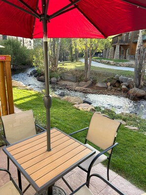 An outdoor patio with a table and chairs under a red umbrella, overlooking a river and a building with a balcony. - On the River, Downtown Estes Park, Close to RMNP Entrance (Estes Park)