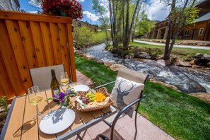 An outdoor dining area with a wooden table, chairs, a basket of fruit, wine glasses, and a bottle. - On the River, Downtown Estes Park, Close to RMNP Entrance (Estes Park)