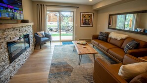 A modern living room with a stone fireplace, a large flat-screen TV, a leather sofa, and a sliding glass door leading to a patio. - On the River, Downtown Estes Park, Close to RMNP Entrance (Estes Park)