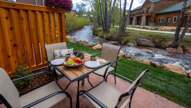 An outdoor patio with a wooden table set for dining, surrounded by chairs, and a view of a river and houses in the background. - On the River, Downtown Estes Park, Close to RMNP Entrance (Estes Park)