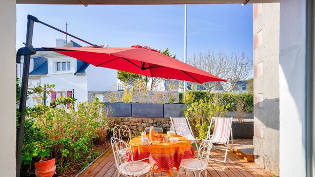 An outdoor patio with a wooden deck, a round table with a red umbrella, white chairs, and a striped towel. - 700m from the beach, charming house for 5 (Penmarch)