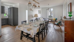 Un comedor con una mesa de madera dispuesta para seis personas, rodeada de sillas. Se observa una zona de cocina con armarios oscuros y una ventana.