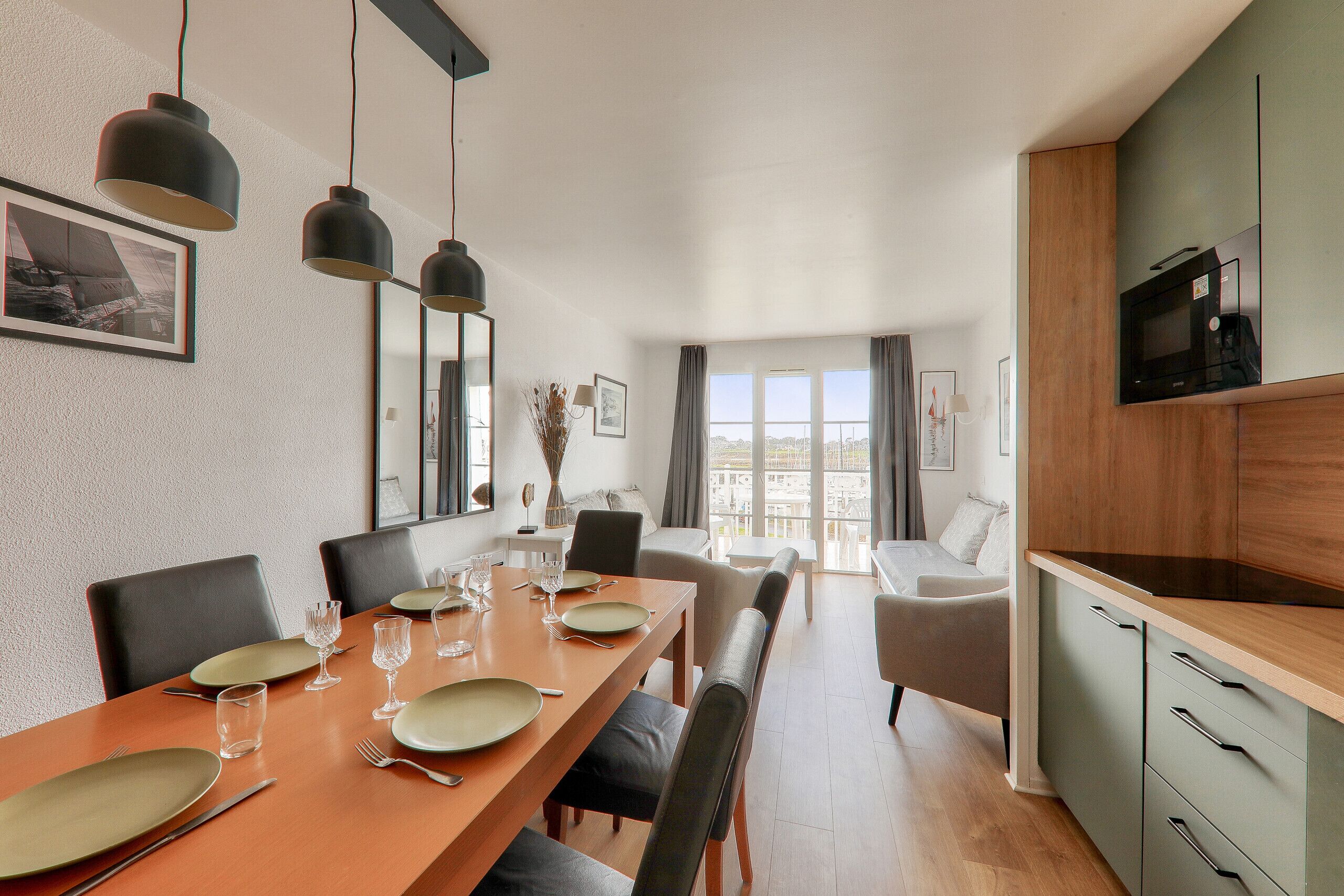 A dining area with a long table set for four, a kitchen with a microwave, and a balcony with a view.