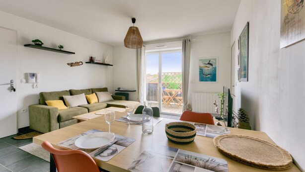 Ein modernes Wohnzimmer mit Esstisch für zwei Personen, einer Couch mit Kissen, einer Pendelleuchte und Blick durch die Glastür.