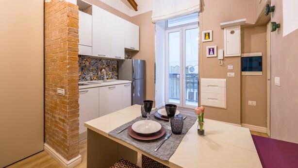 A compact kitchen with white cabinets, a brick accent wall, and a dining area with a table set for two. - Toledo Warm Renovated Studio (Naples)