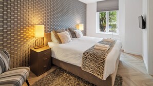 A modern bedroom with a patterned wall, a large bed, a nightstand with a lamp, and a view of trees through a window. - Fully equipped for business trips or cozy meals. (Roosendaal)