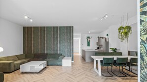 Ein modernes Wohnzimmer mit Fischgrätparkett, einem Ecksofa, einem weißen Couchtisch und einem Essbereich mit einem weißen Tisch und grünen Stühlen.