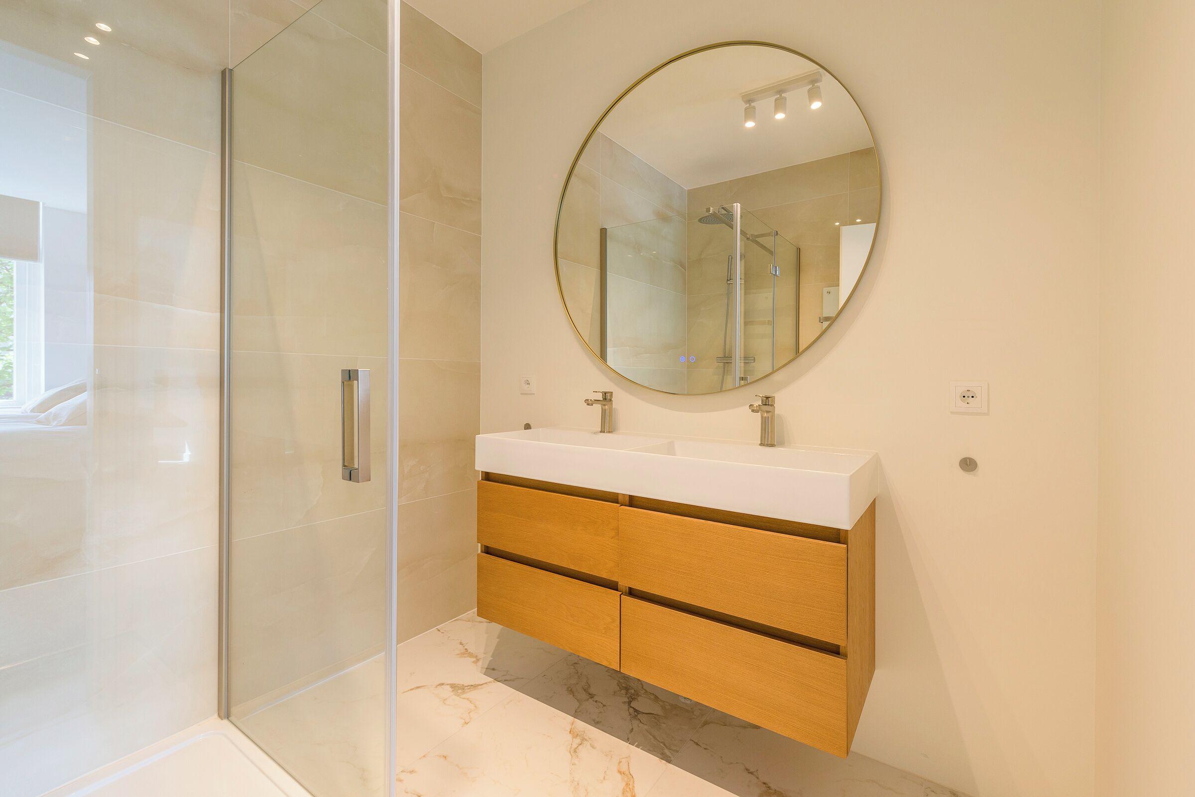 Een moderne badkamer met een glazen douchecabine, een ronde spiegel en een houten wastafelmeubel met een wit aanrechtblad.