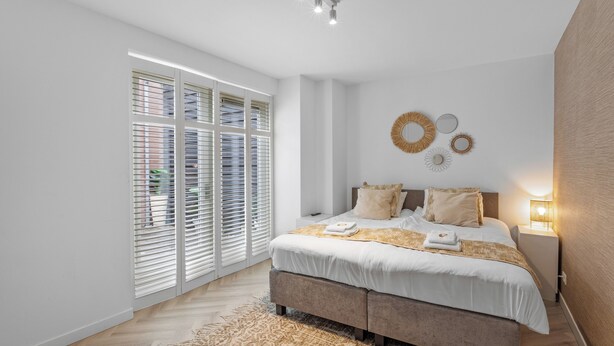 Ein modernes Schlafzimmer mit einem großen Bett, einem Nachttisch mit Lampe und Blick durch raumhohe Fenster.