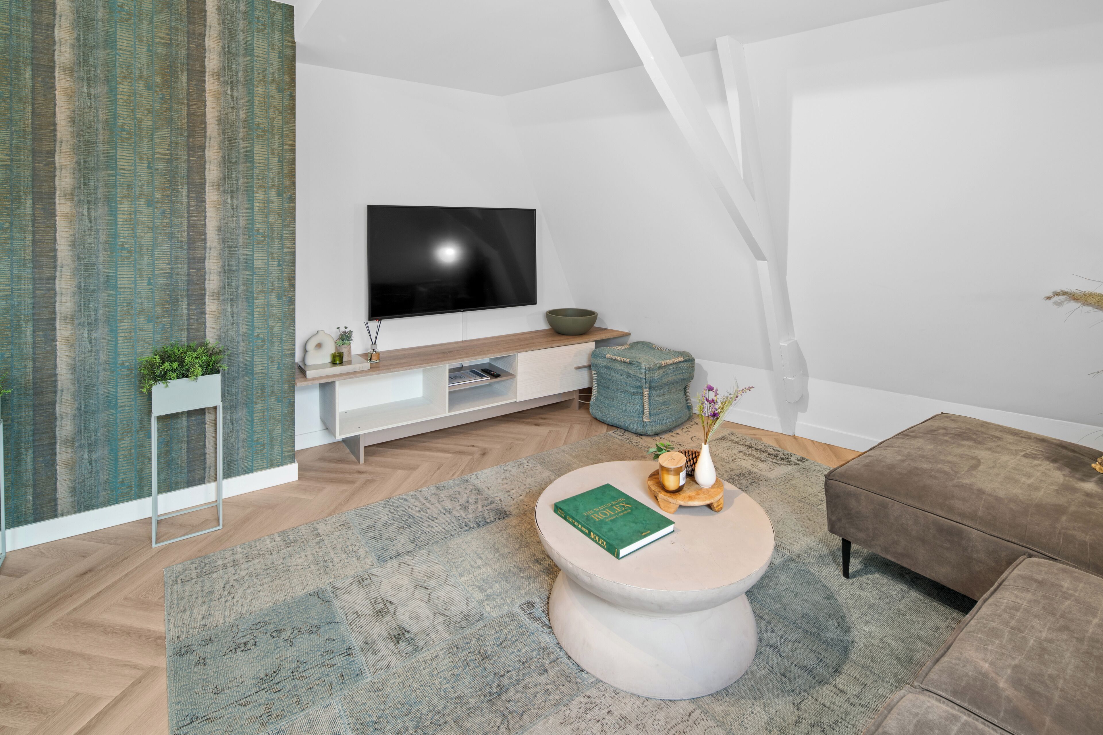 Un salon moderne avec un téléviseur à écran plat, une table basse ronde sur laquelle repose un livre, un canapé gris et un parquet à motifs chevrons.
