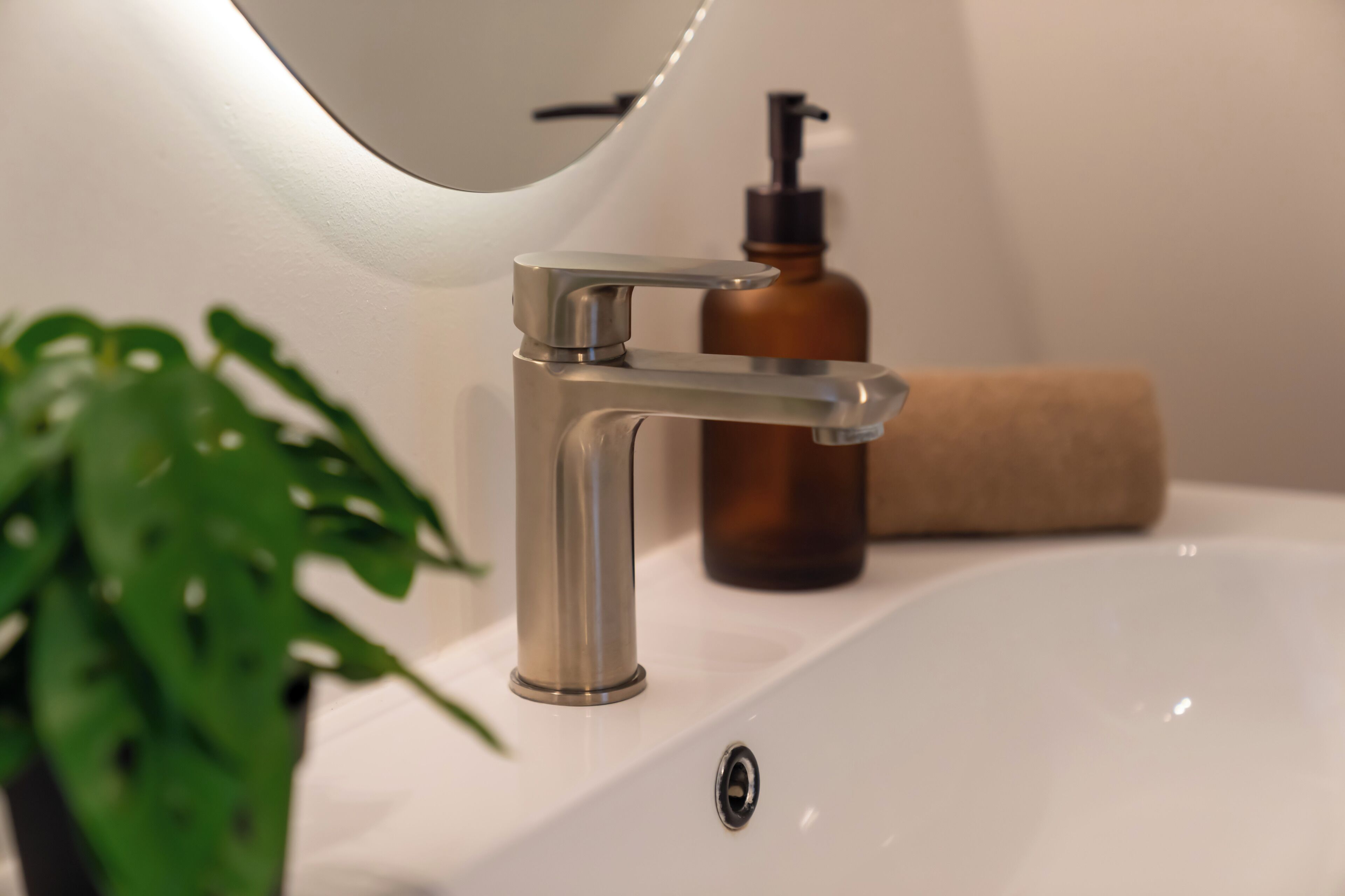 Une salle de bain moderne avec un lavabo blanc, un robinet chromé, un distributeur de savon marron et une plante verte.