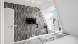 A bedroom with a sloped ceiling, a mounted TV, a bed with neatly folded towels, and a window providing natural light.