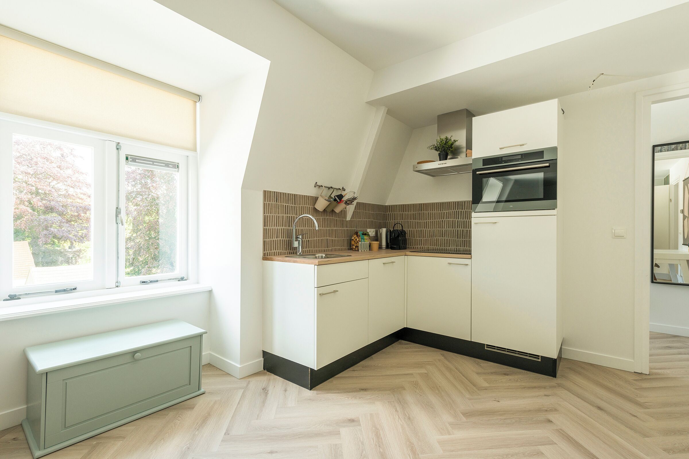 Una cocina moderna con armarios blancos, piso con patrón de espiga, horno empotrado y ventana con persianas.