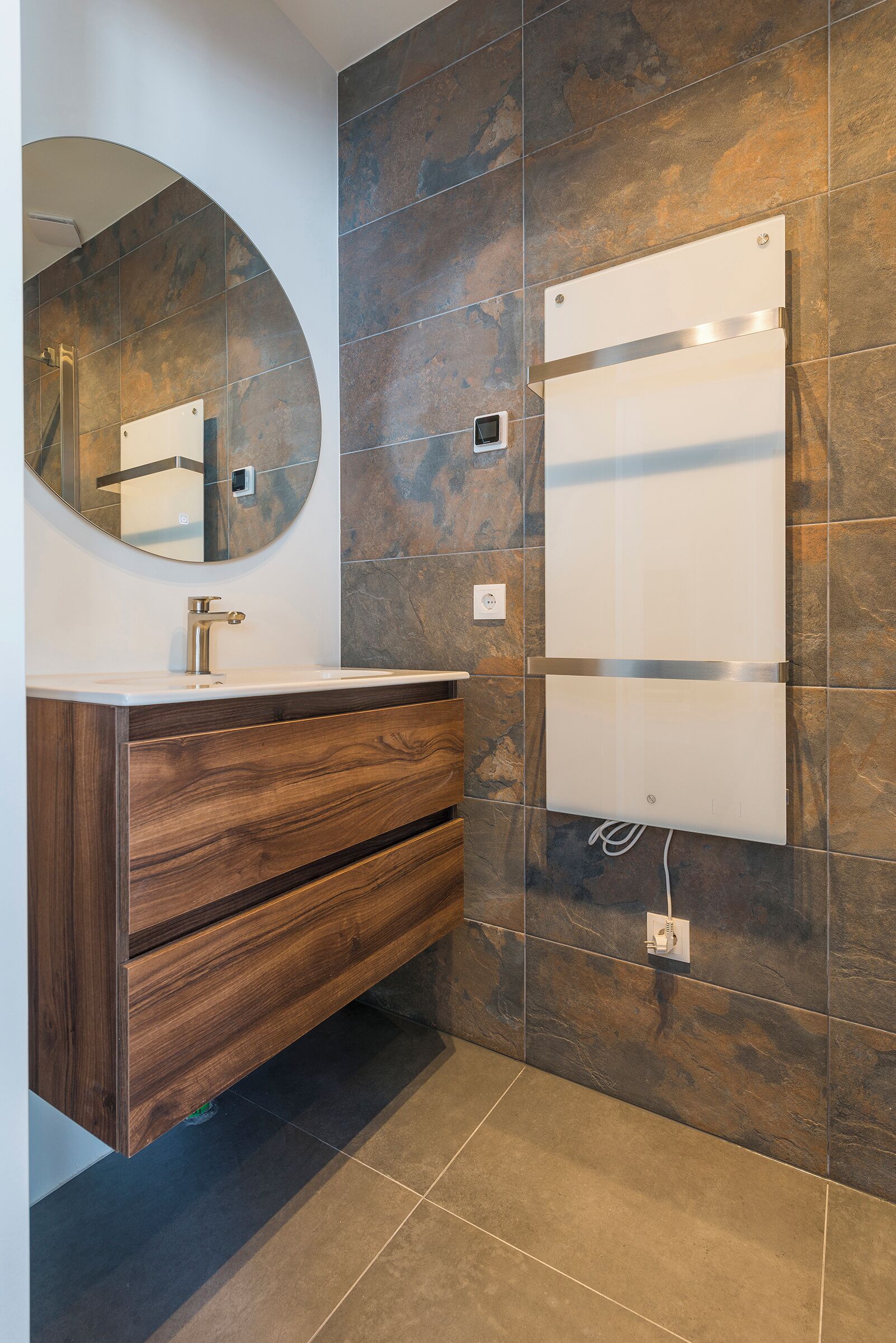 Une salle de bain moderne avec un meuble-lavabo en bois, un miroir rond et un panneau de douche intégré.