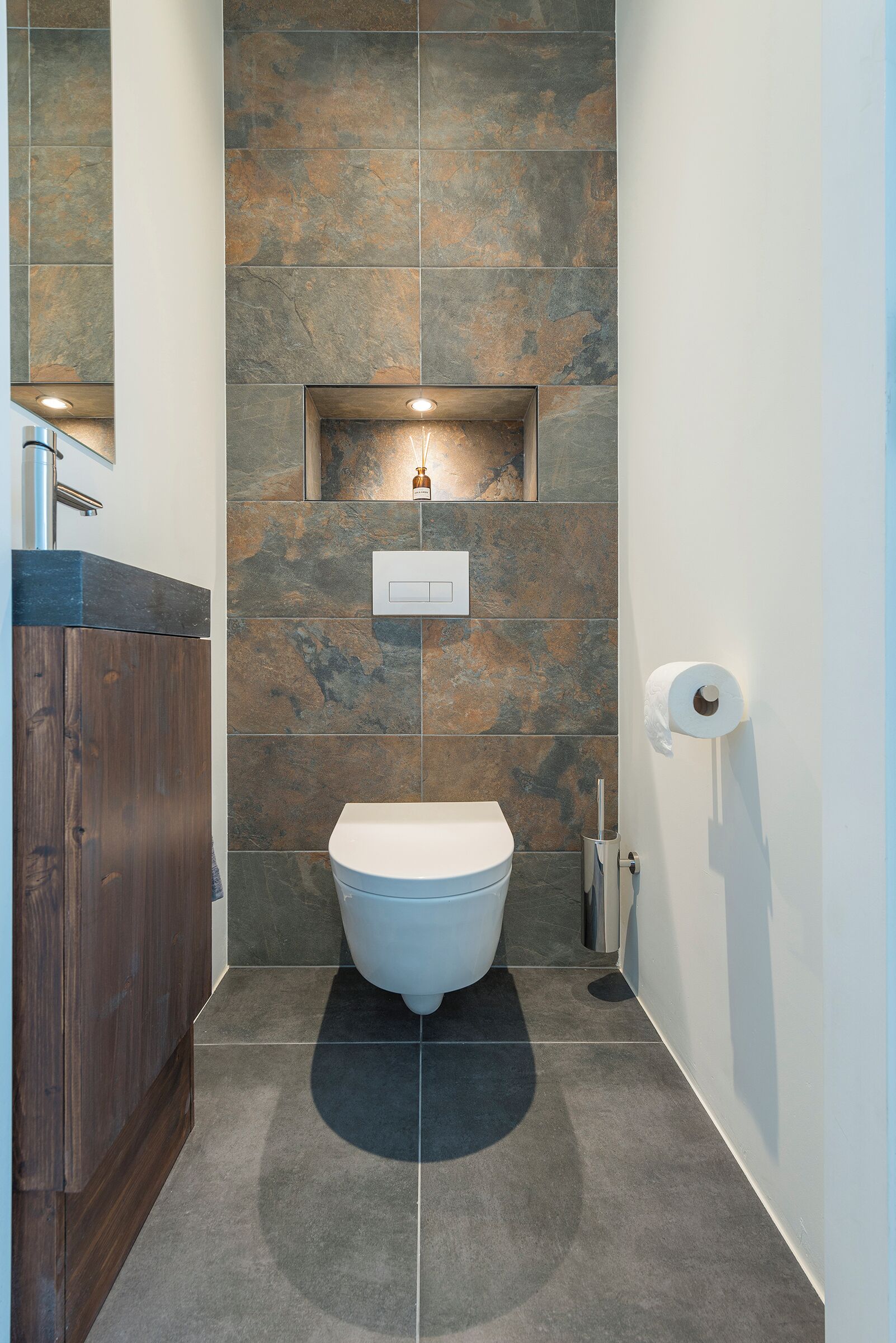 Une salle de bain moderne avec un mur en pierre, un meuble-lavabo en bois, des toilettes blanches et un distributeur de serviettes.