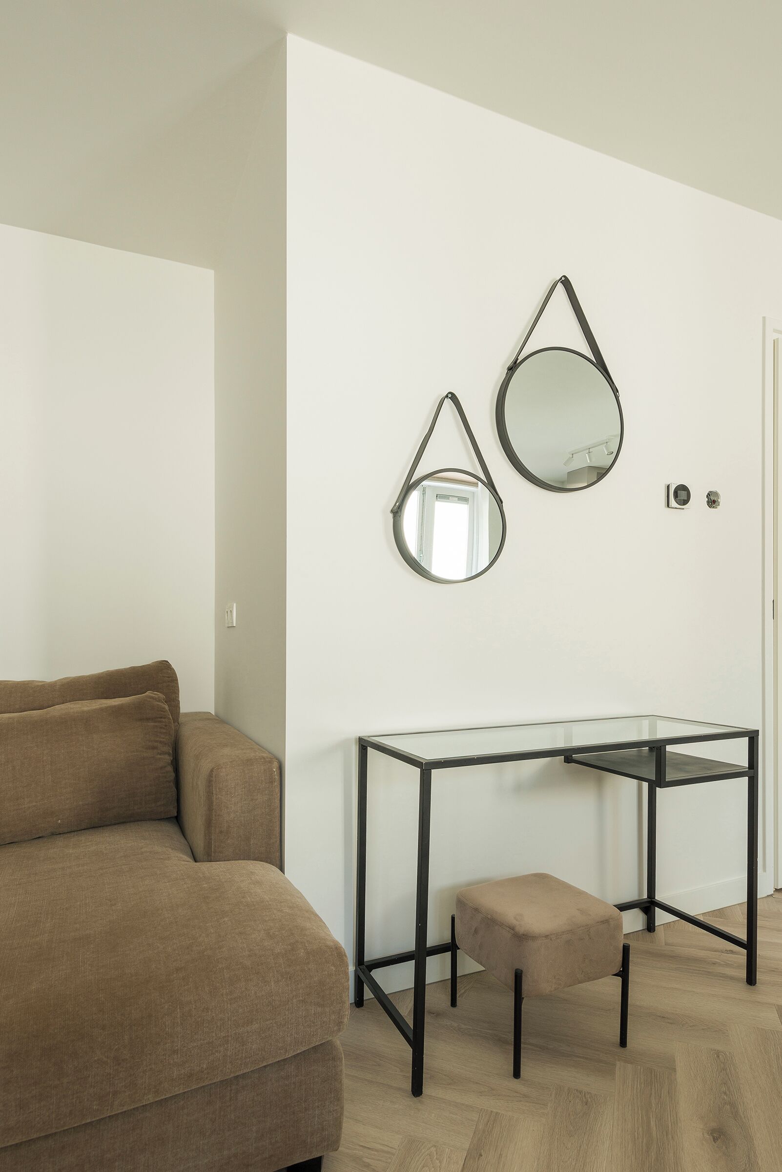 A modern living room with a brown sofa, a glass-top table with a black metal frame, and two wall-mounted mirrors.