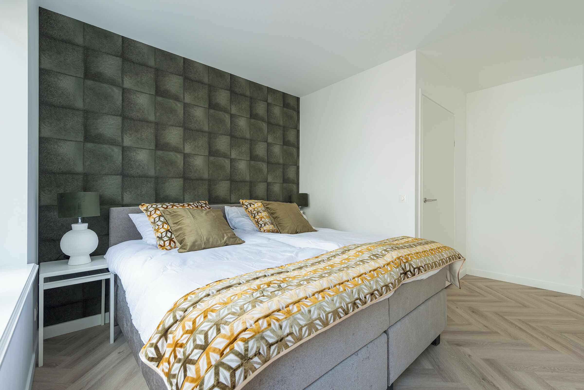 Een moderne slaapkamer met een groot bed, een nachtkastje, een hoofdbord bevestigd aan de muur, en een deur.