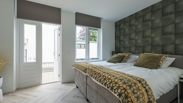 Une chambre à coucher avec un lit, une tête de lit grise, une fenêtre et une porte donnant sur un balcon.