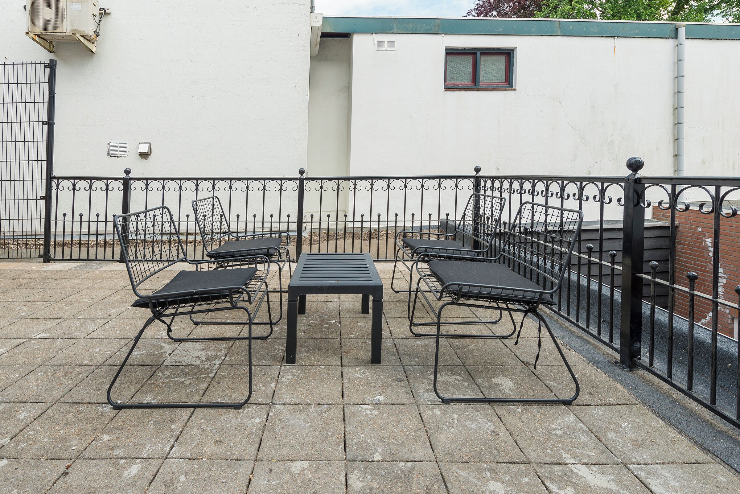 A patio with metal chairs and a table, a black metal fence, and a white building with a window in the background.