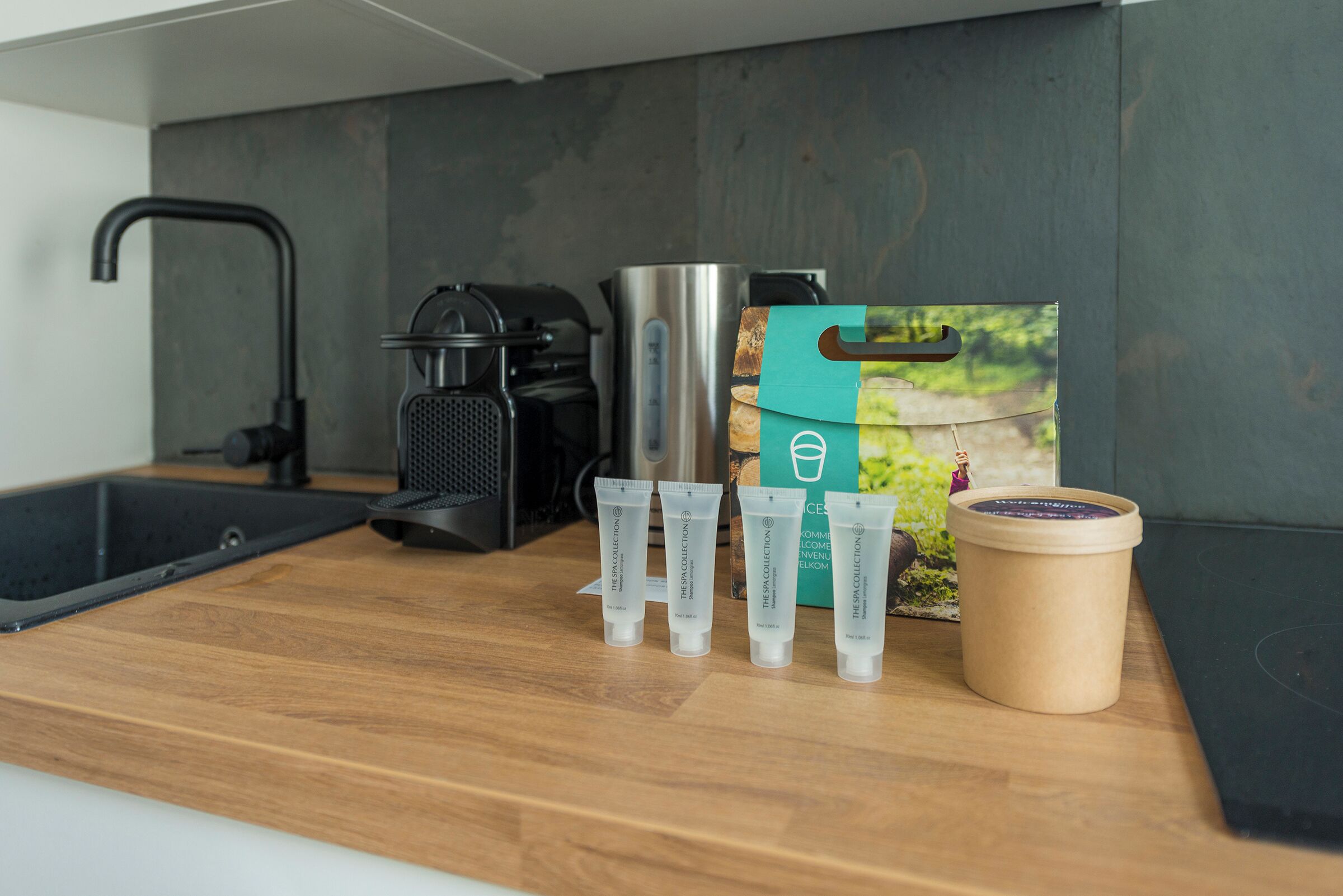 A kitchen counter with a coffee maker, a kettle, and a box of The Space Collection skincare products.