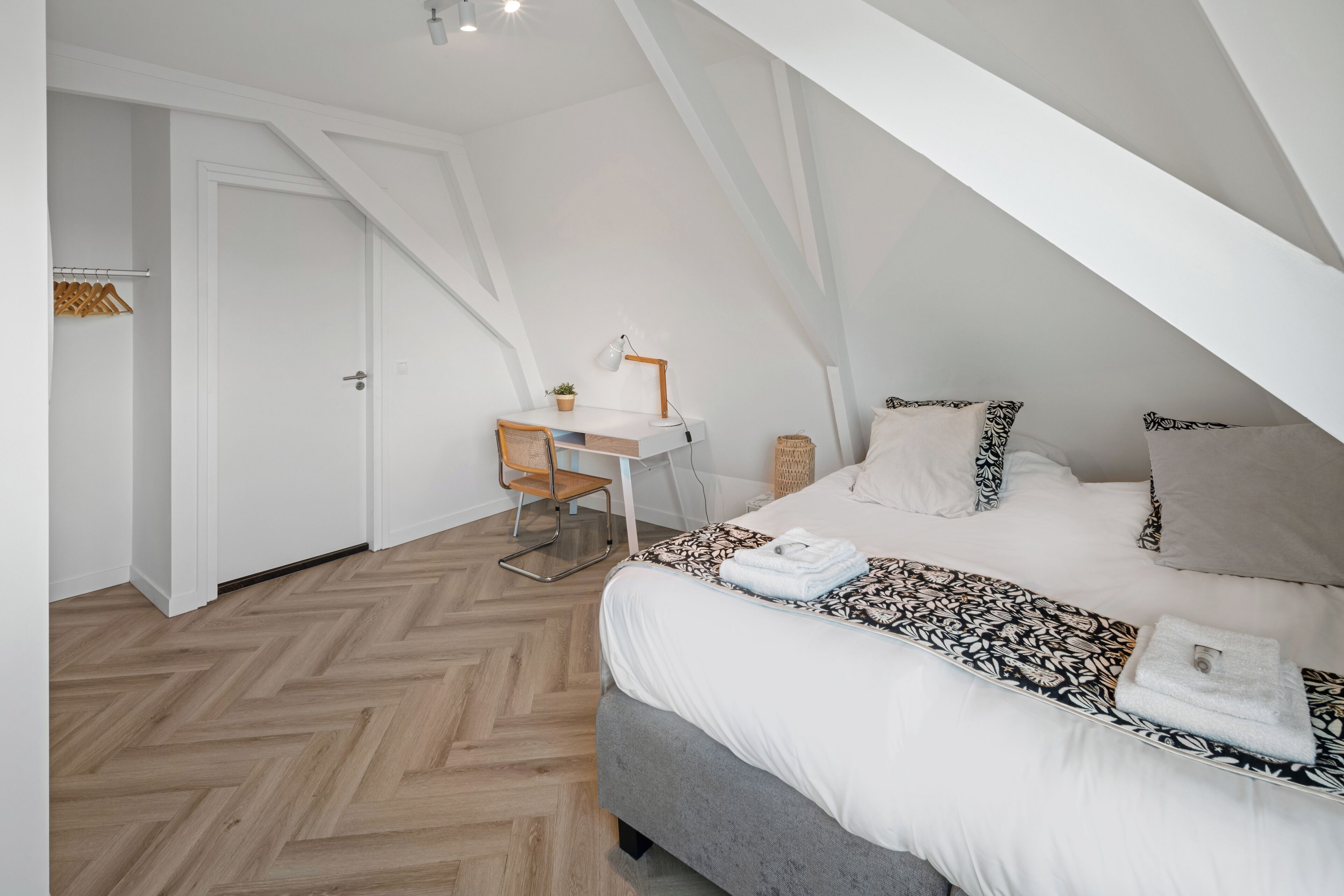 A bedroom with a herringbone floor, a bed with patterned bedding, a desk with a lamp, and a chair.