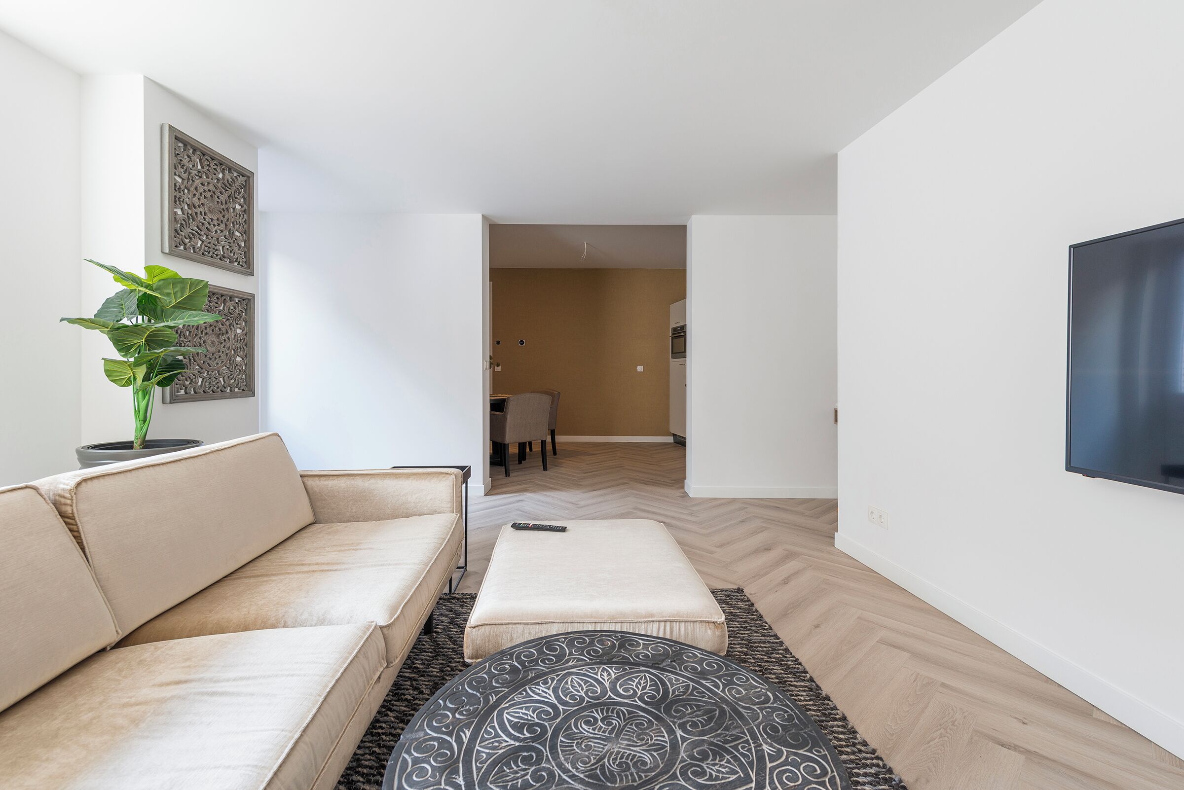 A modern living room with a beige sofa, a patterned ottoman, and a potted plant.