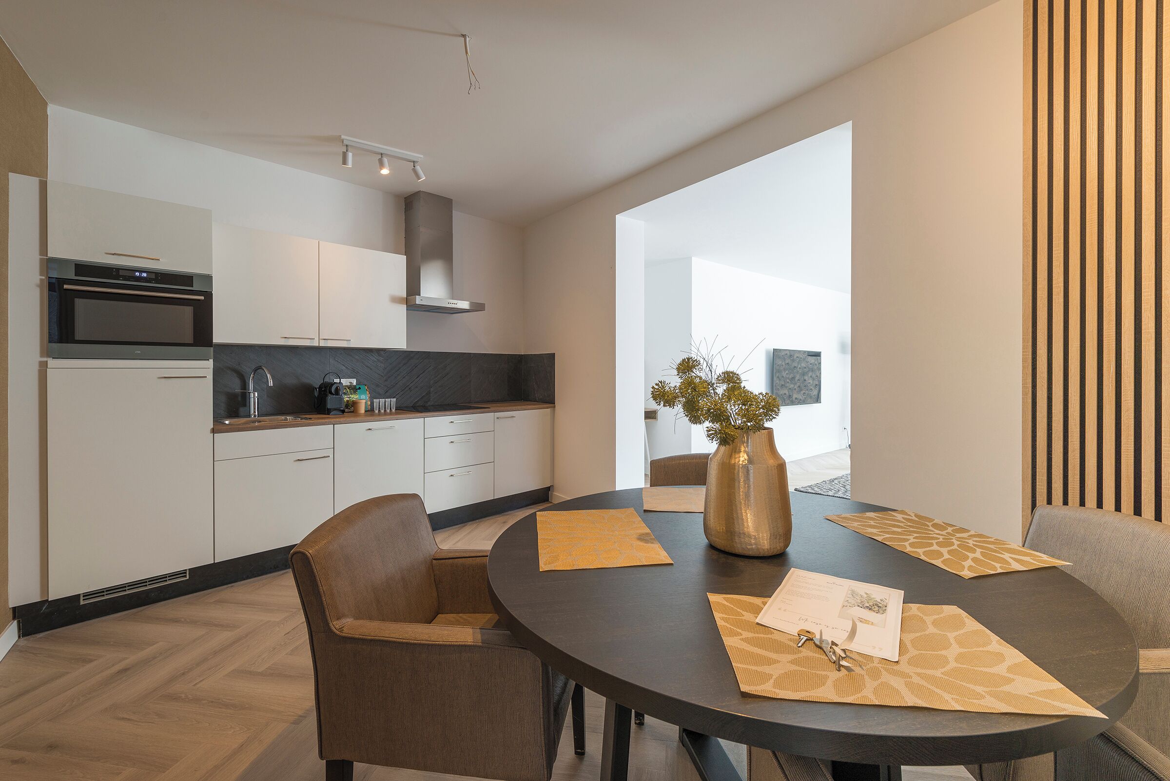 Een moderne keuken met witte kasten, een roestvrijstalen oven en een zwart aanrecht. Er is een zithoek met een ronde tafel, stoelen en een vaas met bloemen.