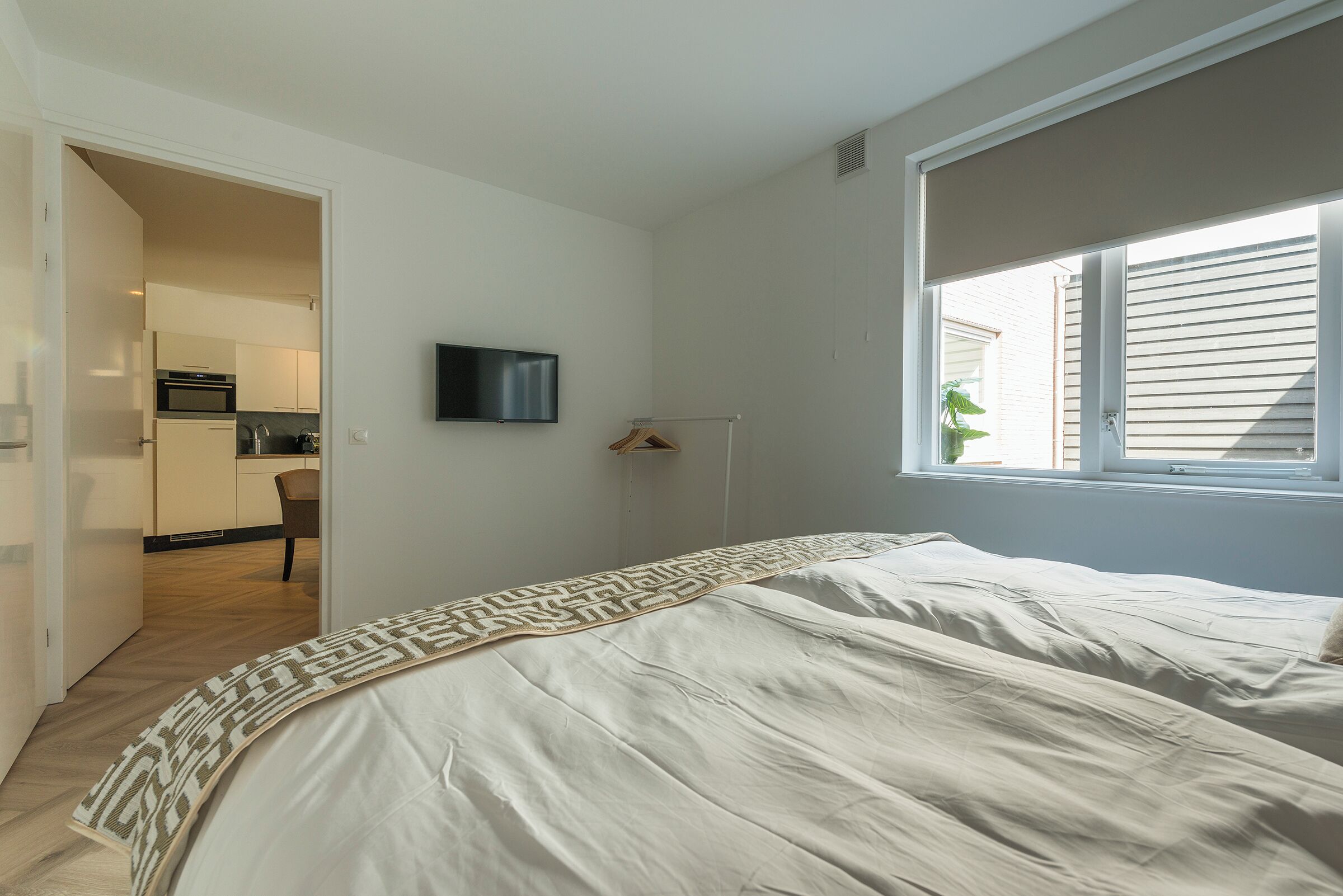 A bedroom with a bed, a TV, a window, and a door leading to a kitchen.