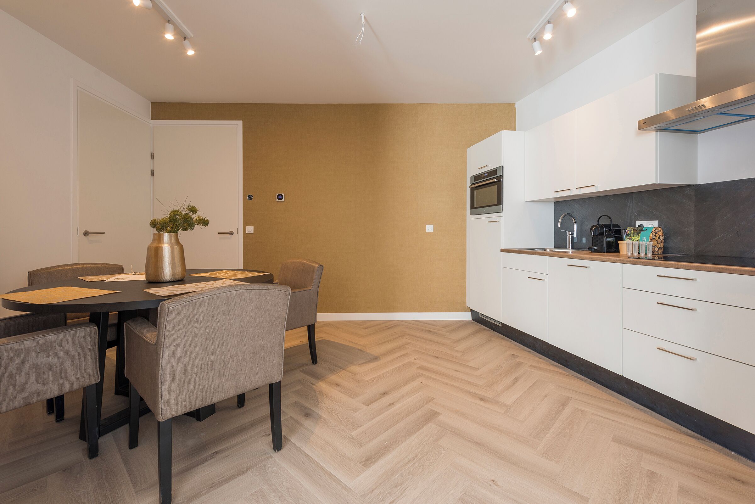 Uma cozinha moderna com armários brancos, piso de madeira em padrão herringbone e uma área de refeições com mesa e cadeiras.
