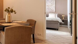 Moderni hotellihuone, jossa on ruokailutila, harmaalla päätyosalla varustettu sänky ja suuri, abstrakti taulu seinällä.