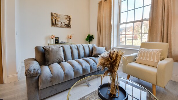 Un salon moderne avec un canapé gris, un fauteuil beige, une table basse en verre et une grande fenêtre avec des rideaux.