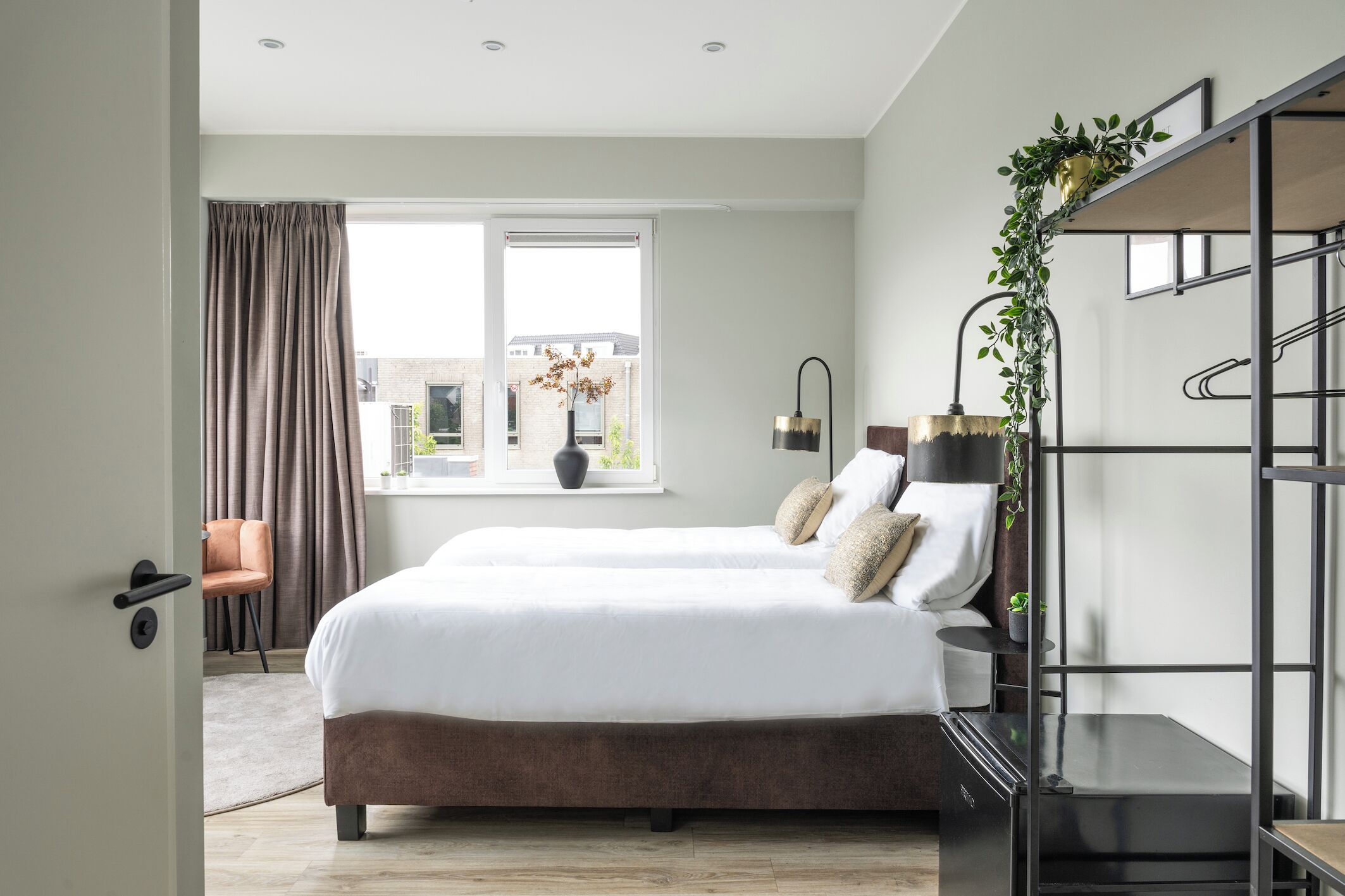 Bright and modern bedroom with twin beds, white linens, gold-accented lamps, greenery, open shelving, neutral tones, and a large window for natural light.