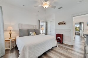 A bedroom with a bed, bedside lamps, a ceiling fan, a nightstand, and a small red table. - Cottages at Romar C1 - Gulf Side Cottage! (Orange Beach)