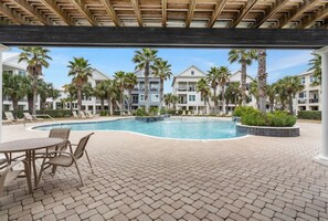A pool area with lounge chairs and a table, surrounded by palm trees and multi-story buildings. - Cottages at Romar C1 - Gulf Side Cottage! (Orange Beach)