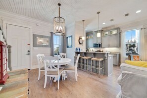 A modern kitchen with white cabinets, a central island, and a dining area with white chairs. - Cottages at Romar C1 - Gulf Side Cottage! (Orange Beach)