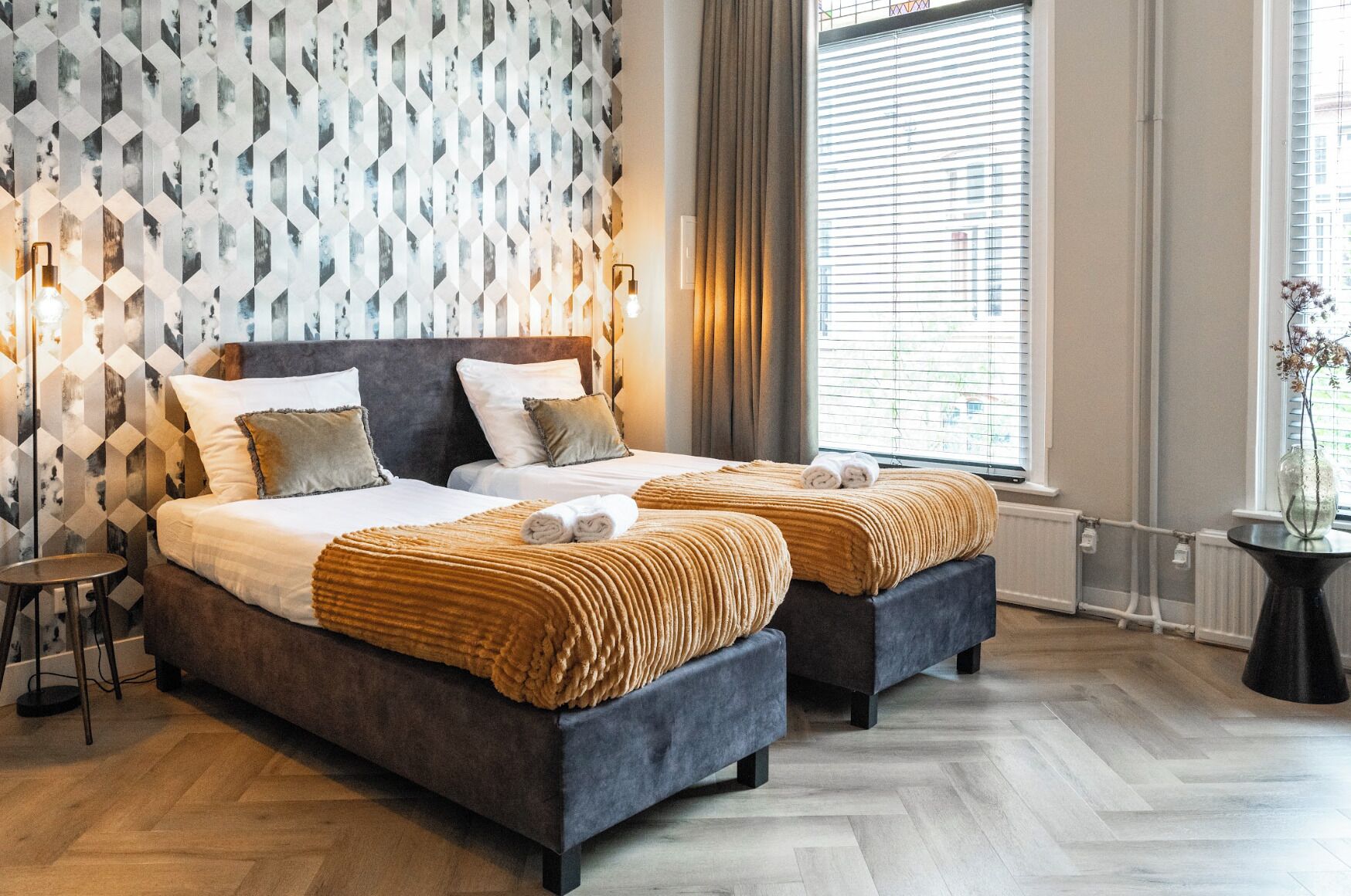 Habitación de hotel con dos camas, un papel tapiz estampado, una ventana con persianas y una mesita con un florero.