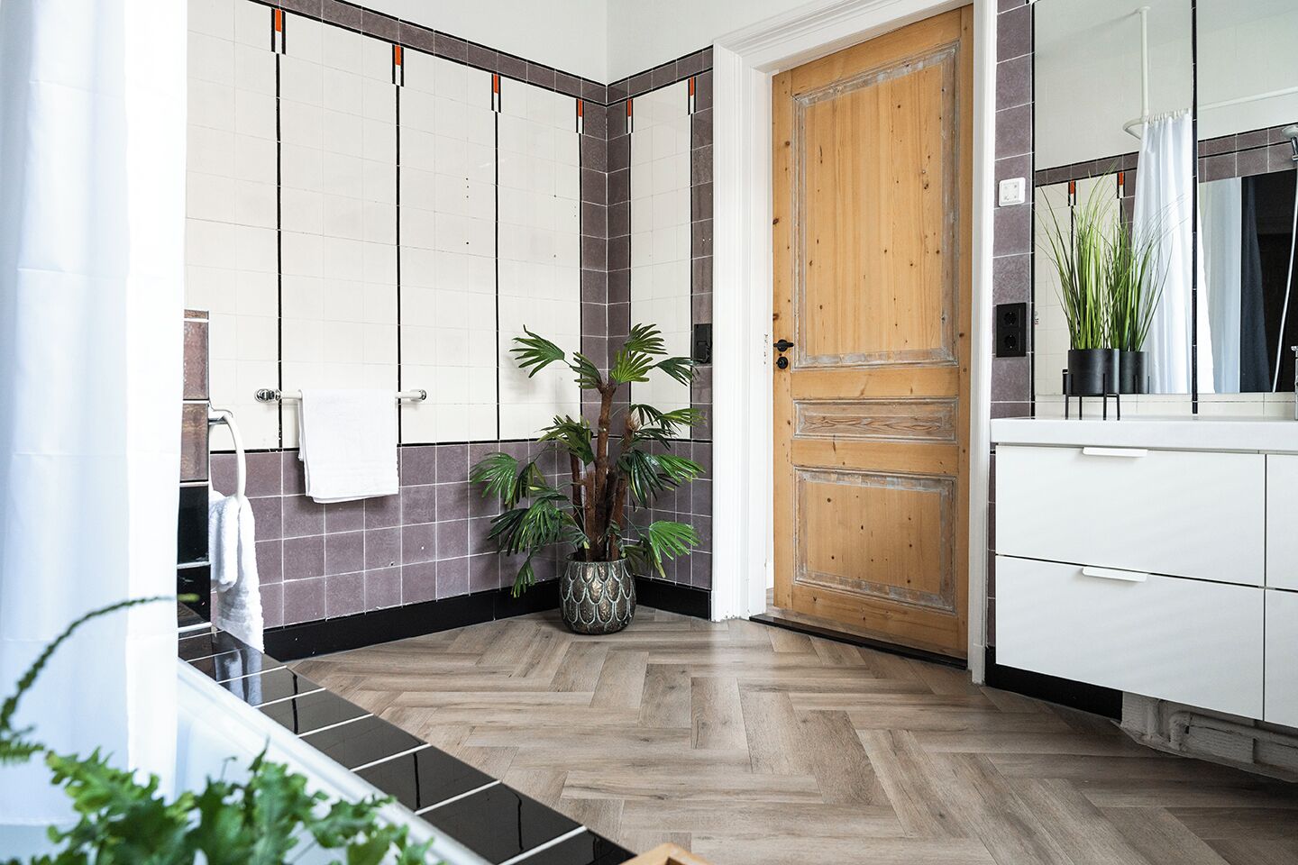 A bathroom with a herringbone wood floor, white cabinets, a wooden door, and a potted plant.