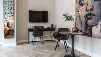 Un salon moderne avec un parquet en chevrons, un téléviseur à écran plat fixé au mur, une table à manger avec deux chaises et un grand tableau abstrait.