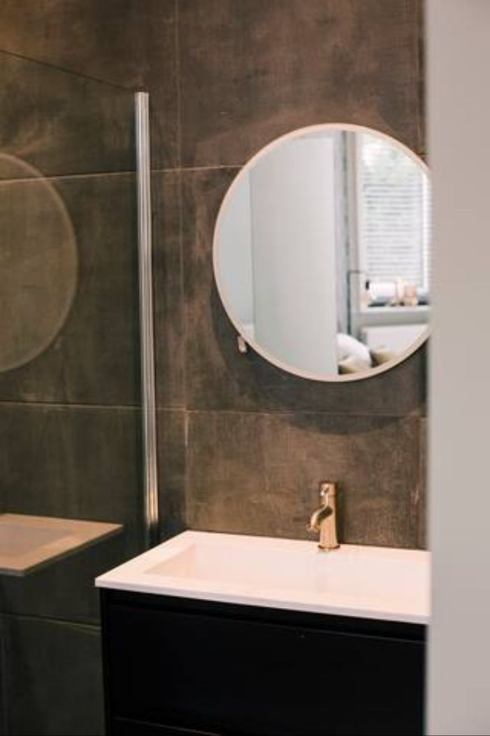 Un baño con pared de azulejos oscuros, un espejo redondo, un lavabo blanco con grifo dorado y un panel de vidrio vertical.