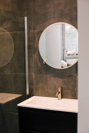 Un bany amb una paret revestida amb rajoles fosques, un mirall rodó, un lavabo blanc i una separació de vidre vertical.