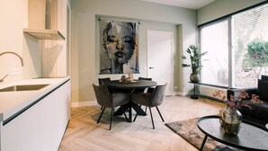 Een moderne keuken met een eethoek, een groot abstract schilderij en uitzicht op groen door het raam.