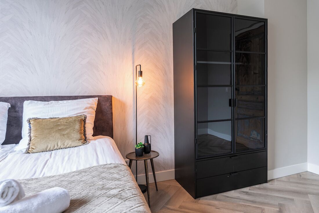 Un dormitorio con una cama, una mesita de noche con lámpara y un amplio armario negro.